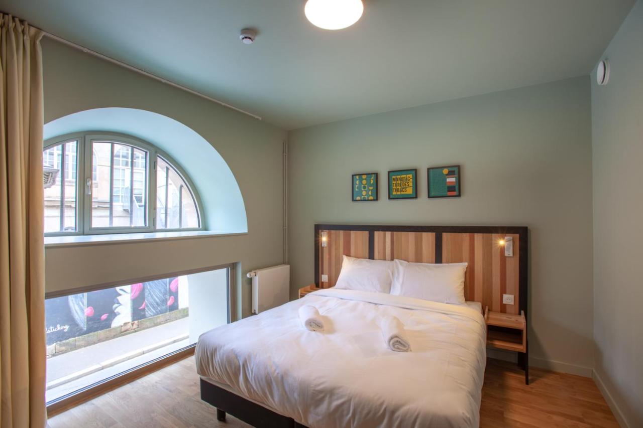 The People - Strasbourg Hostel Εξωτερικό φωτογραφία The photo shows a cozy bedroom with a light green wall. It features a double bed with a white comforter, and there are bedside tables on either side of the bed. Above the bed, there are three framed pieces of artwork. A large arched window provides n