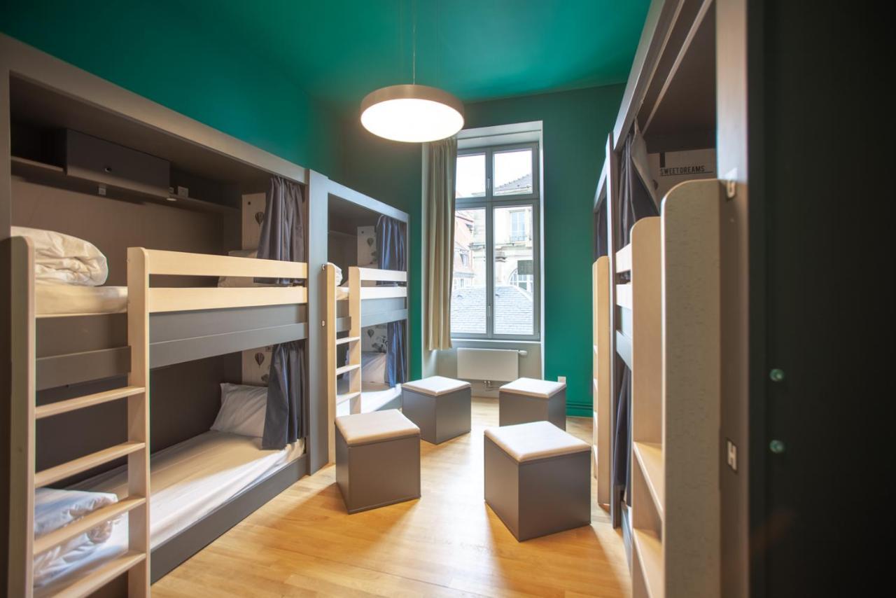 The People - Strasbourg Hostel Εξωτερικό φωτογραφία The photo shows a modern hostel-style room featuring two sets of bunk beds along one wall. The beds have a simple, sleek design and are outfitted with pillows and blankets. In the center of the room, there are several small gray stools or tables, pro