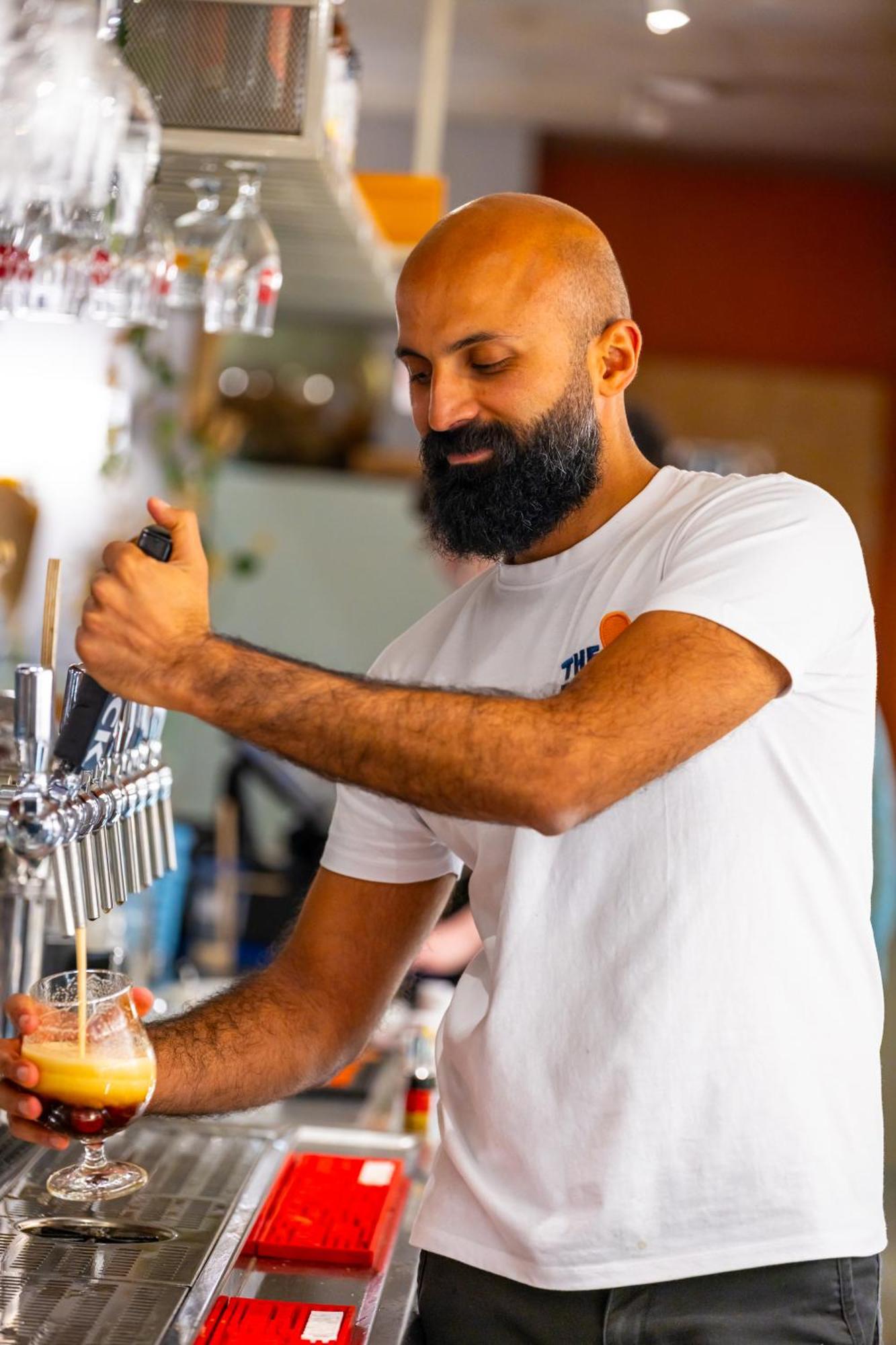 The People - Strasbourg Hostel Εξωτερικό φωτογραφία The photo shows a man with a beard wearing a white T-shirt, working behind a bar. He is actively pouring a drink from a tap, using one hand to grip the handle. The background suggests a lively bar environment, with various items and people slightly b