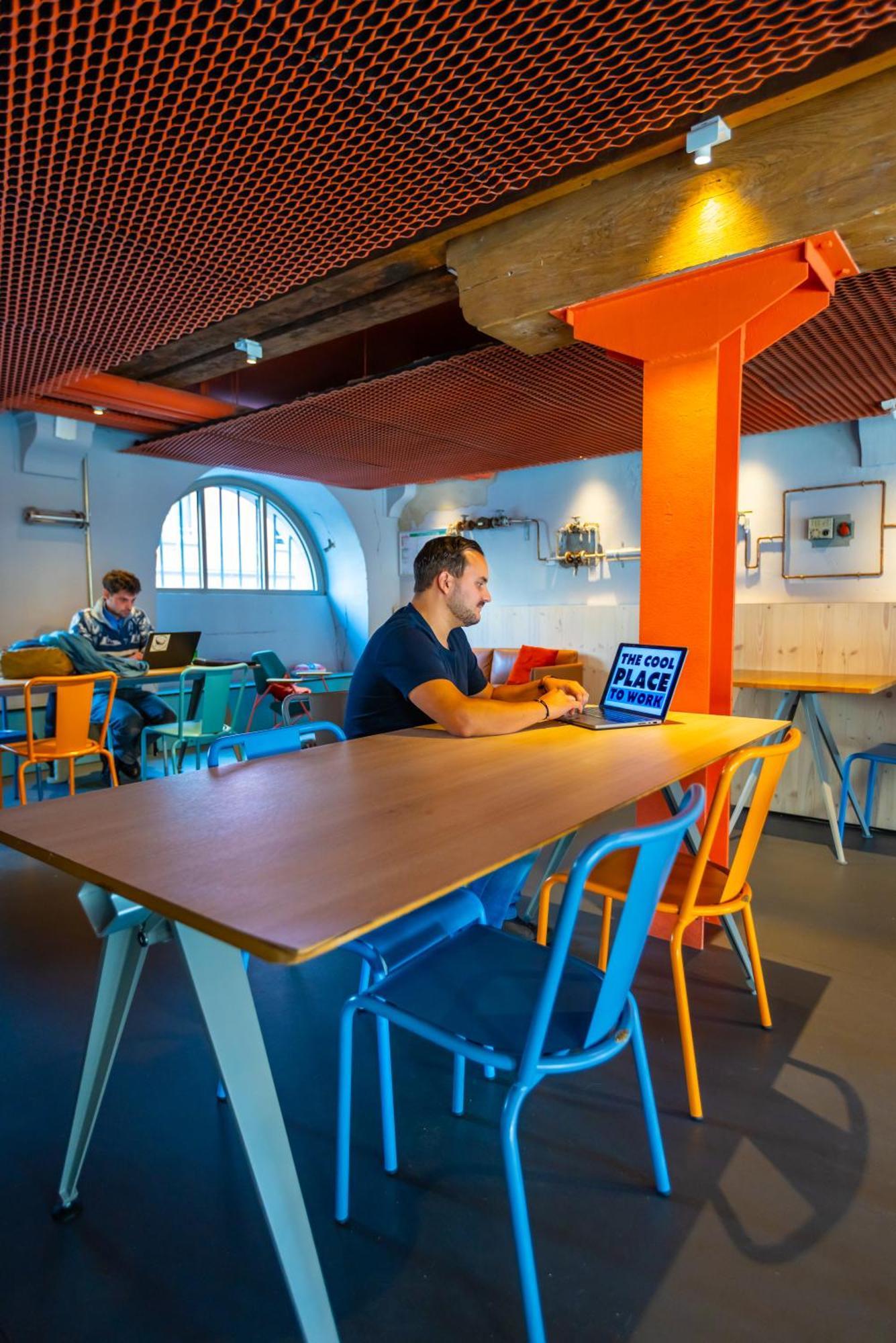 The People - Strasbourg Hostel Εξωτερικό φωτογραφία The photo depicts a modern and vibrant indoor space designed for work or socializing. In the foreground, a man is seated at a long, light wooden table, focused on his laptop. He is surrounded by colorful chairs in shades of blue, orange, and yellow. 