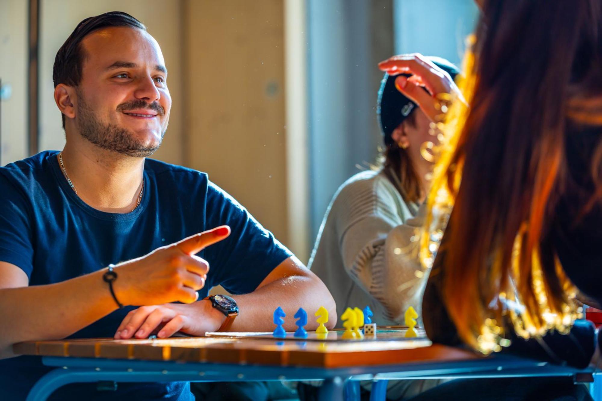 The People - Strasbourg Hostel Εξωτερικό φωτογραφία The photo shows a man sitting at a table, engaging in a conversation with a woman who is partially visible. The man is smiling and pointing, likely animatedly discussing something. On the table in front of them, there are colorful game pieces arrange