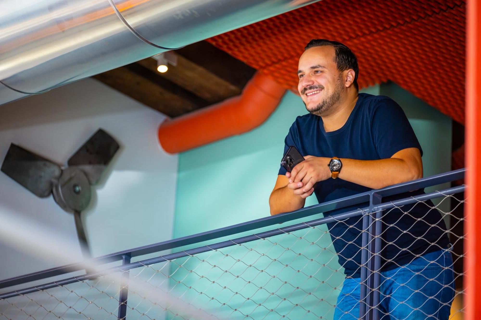 The People - Strasbourg Hostel Εξωτερικό φωτογραφία The photo shows a man standing on a balcony or a railing in a modern indoor setting. He has a relaxed posture, leaning slightly forward with a smile on his face. He is wearing a dark blue T-shirt and a watch on his left wrist. The background features