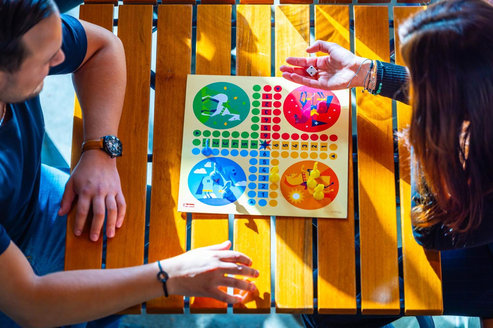 The People - Strasbourg Hostel Εξωτερικό φωτογραφία The photo shows two people sitting at a wooden table, engaged in a board game. The game board features four colored quadrants—red, yellow, blue, and green—each with illustrations and a grid of colored pieces. One person is rolling a die, while the ot