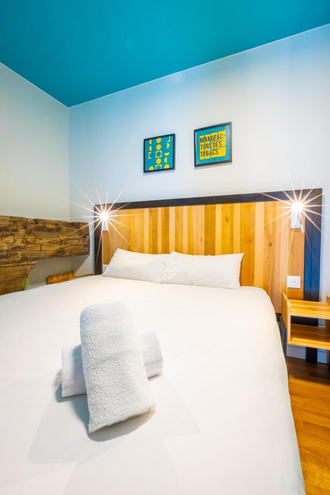 The People - Strasbourg Hostel Εξωτερικό φωτογραφία The photo shows a cozy bedroom featuring a double bed with a wooden headboard. The bed is neatly made with white bedding and a rolled-up towel placed on it. There are two small bedside lights at the head of the bed. The walls are painted in a light b