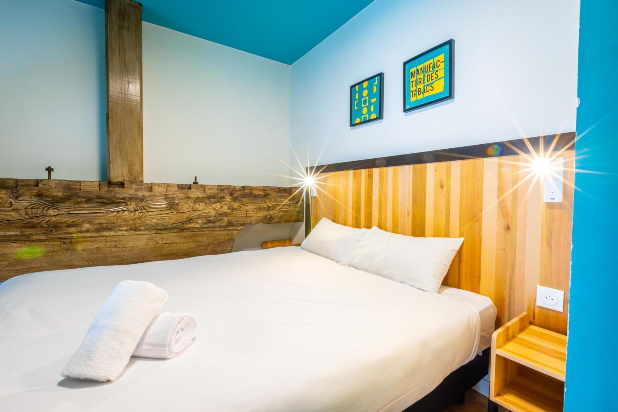 The People - Strasbourg Hostel Εξωτερικό φωτογραφία The photo shows a cozy hotel room featuring a double bed with white linens and two pillows. The headboard of the bed has a wooden design with a light wood finish, complemented by a turquoise wall above. There are two framed pieces of art on the wall,
