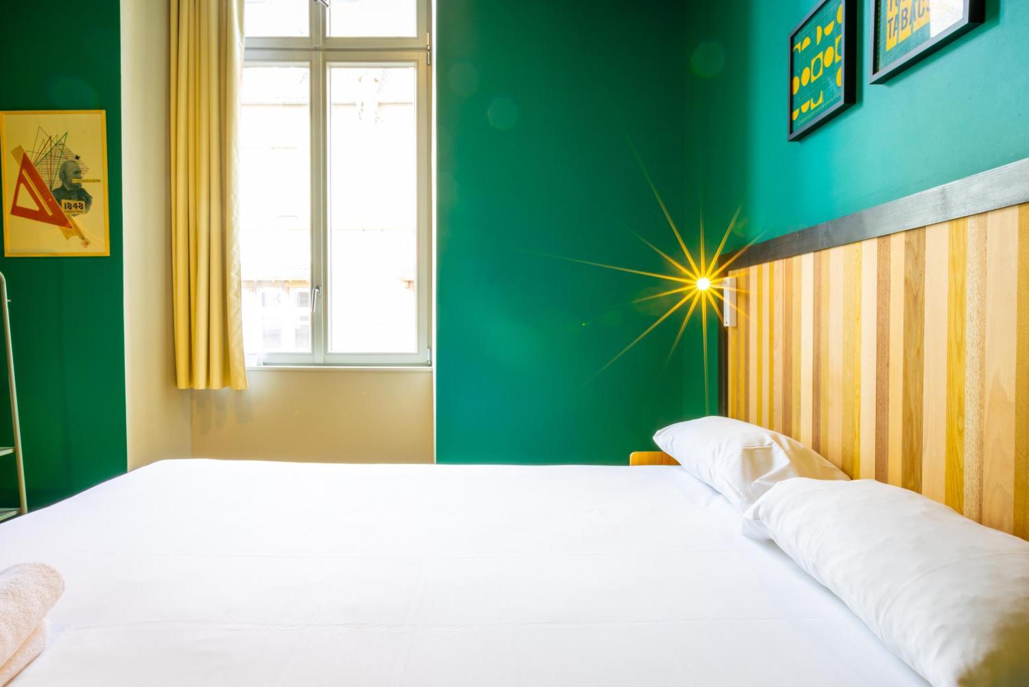 The People - Strasbourg Hostel Εξωτερικό φωτογραφία The photo shows a modern bedroom with a vibrant green wall. The bed is positioned in the foreground with a simple white bedspread and two white pillows. A wooden headboard adds a warm touch. To the side, there is a large window with sheer yellow curt