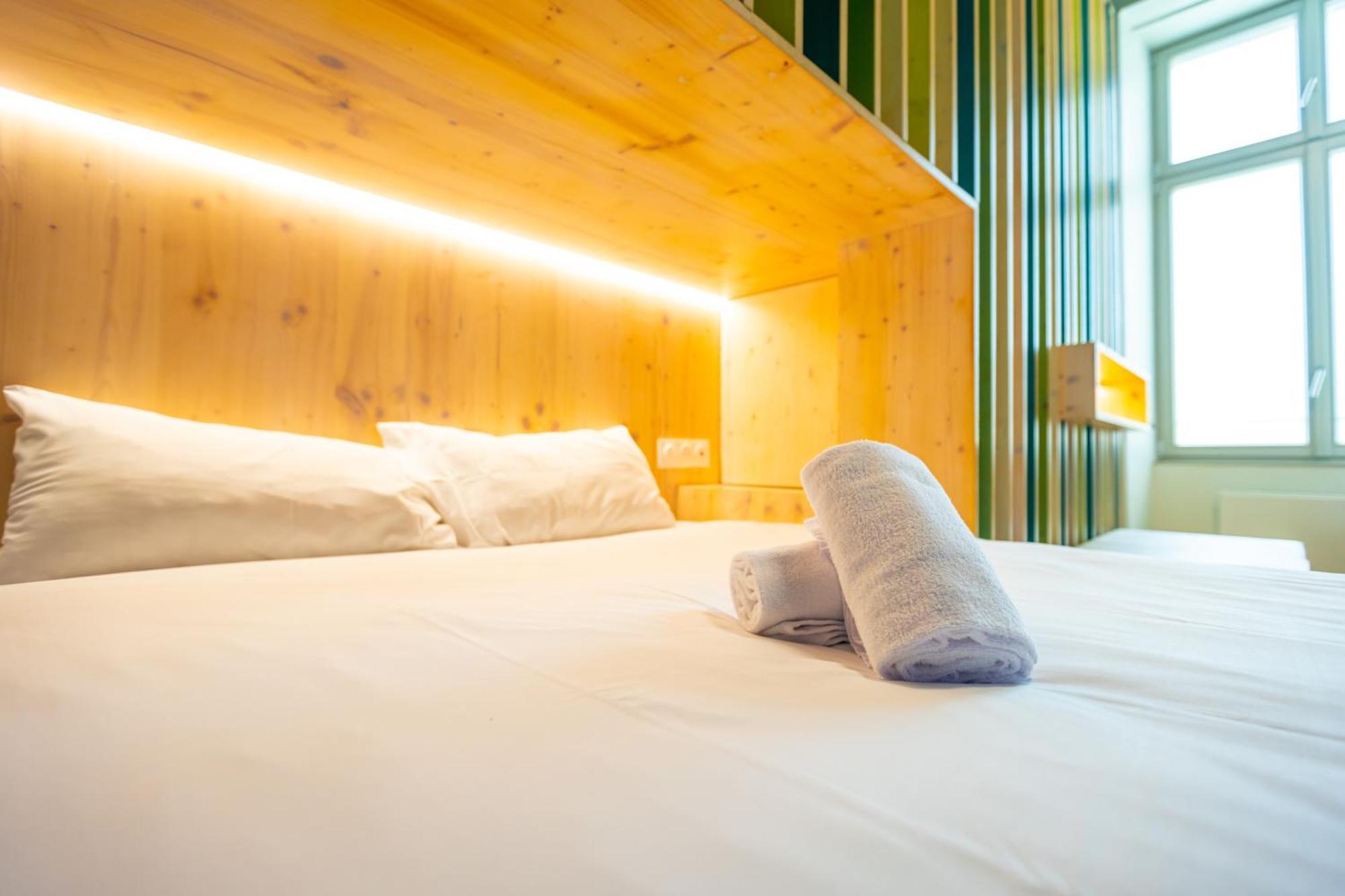 The People - Strasbourg Hostel Εξωτερικό φωτογραφία The photo shows a cozy bedroom setting featuring a bed with white bedding. The headboard of the bed is made of light-colored wood and has integrated lighting that provides a warm glow. Two rolled towels are neatly placed on the bed. The room features
