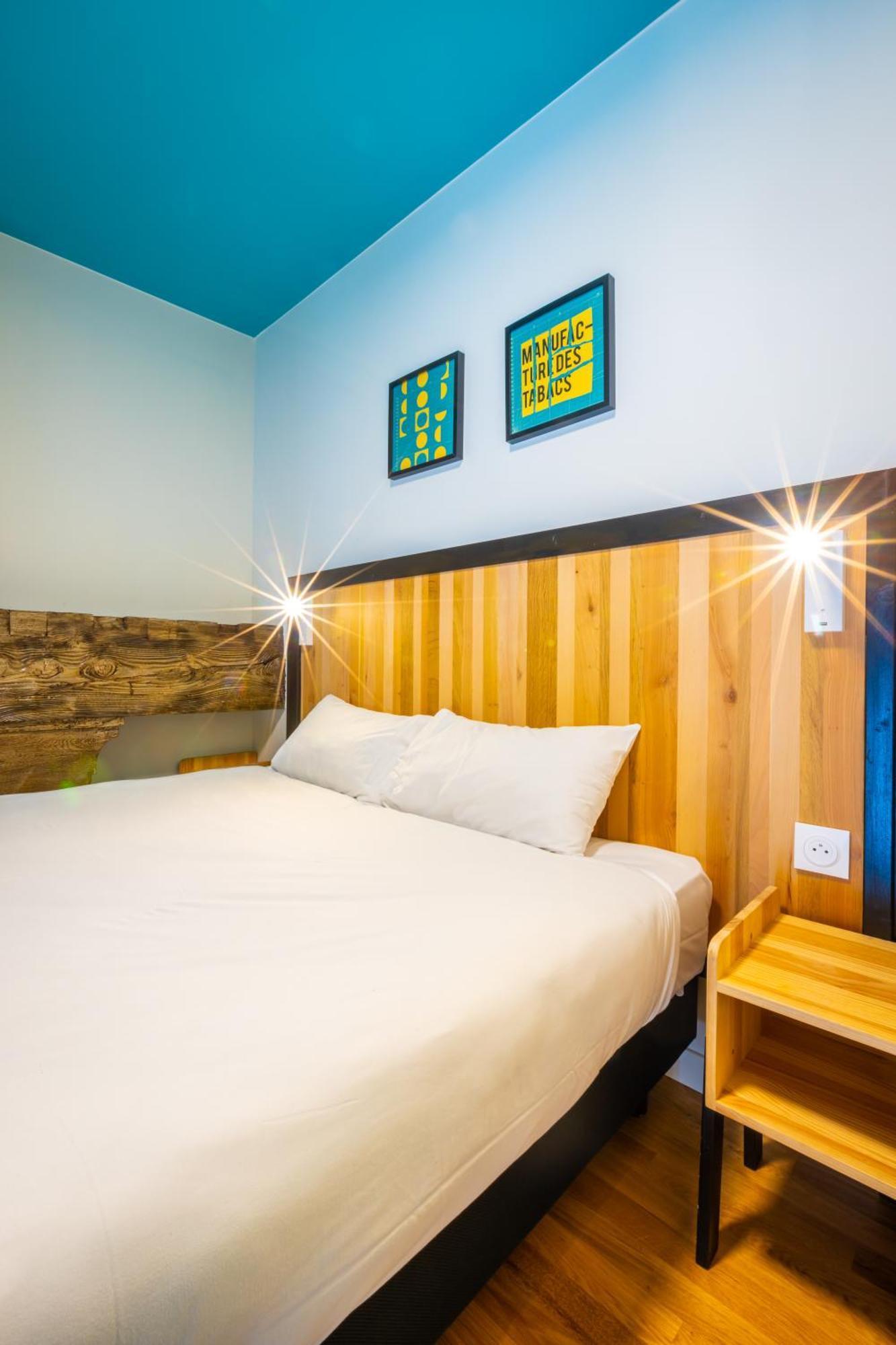 The People - Strasbourg Hostel Εξωτερικό φωτογραφία The photo shows a cozy bedroom setting. It features a bed with a white bedspread and two pillows. The headboard is made of wooden slats, combining light and dark wood tones. On the wall above the bed, there are two framed artworks, likely colorful an
