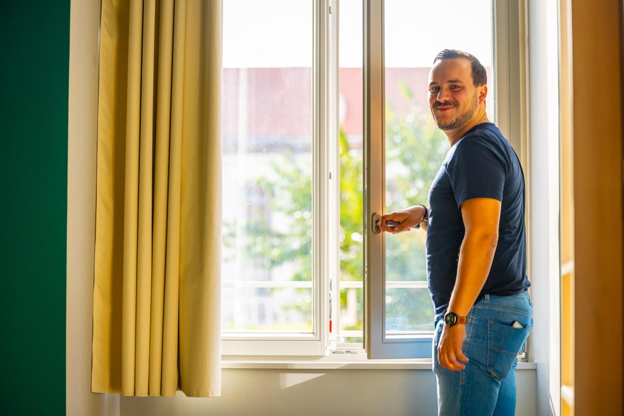 The People - Strasbourg Hostel Εξωτερικό φωτογραφία The photo shows a man standing next to a window, smiling as he opens it. The window has vertical slats and is positioned within a well-lit room, with sunlight streaming in. There are light-colored curtains hanging beside the window. In the background