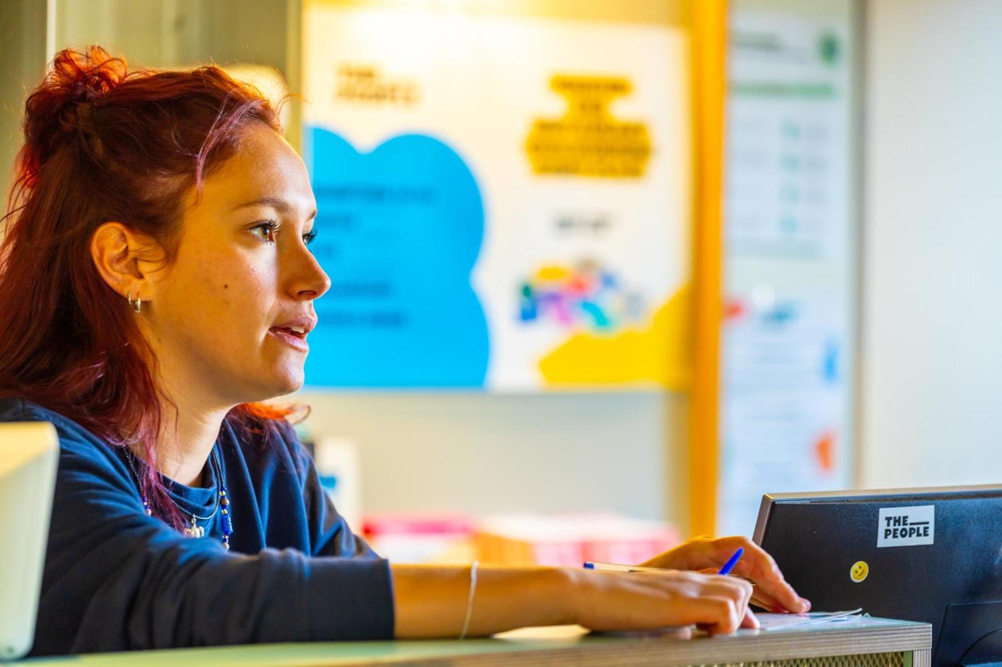 The People - Strasbourg Hostel Εξωτερικό φωτογραφία The photo shows a woman with red hair, sitting at a reception or counter. She appears to be engaged in conversation or focused on something in front of her. In the background, there are colorful posters or signs, suggesting a welcoming or community-o