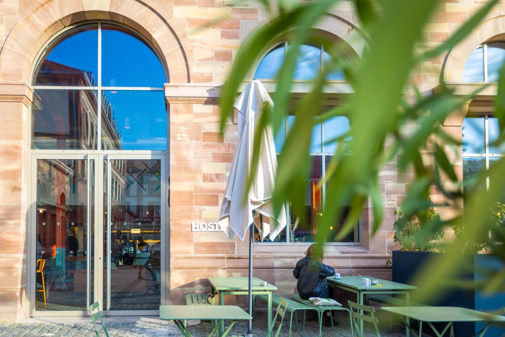 The People - Strasbourg Hostel Εξωτερικό φωτογραφία The photo shows a café or restaurant with a modern outdoor seating area. There are green tables and chairs set outside, with one umbrella providing shade. The architecture features large arched windows that reflect blue skies and nearby buildings. In