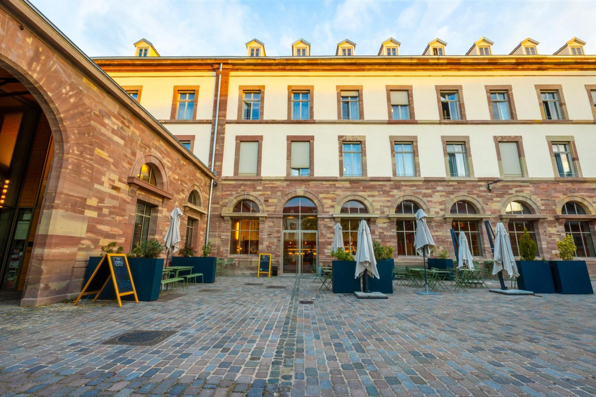 The People - Strasbourg Hostel Εξωτερικό φωτογραφία The photo shows a picturesque courtyard area of a building with a historical and architectural style. The structure features a series of arched windows and an extended façade. In the foreground, there are outdoor seating arrangements, including table