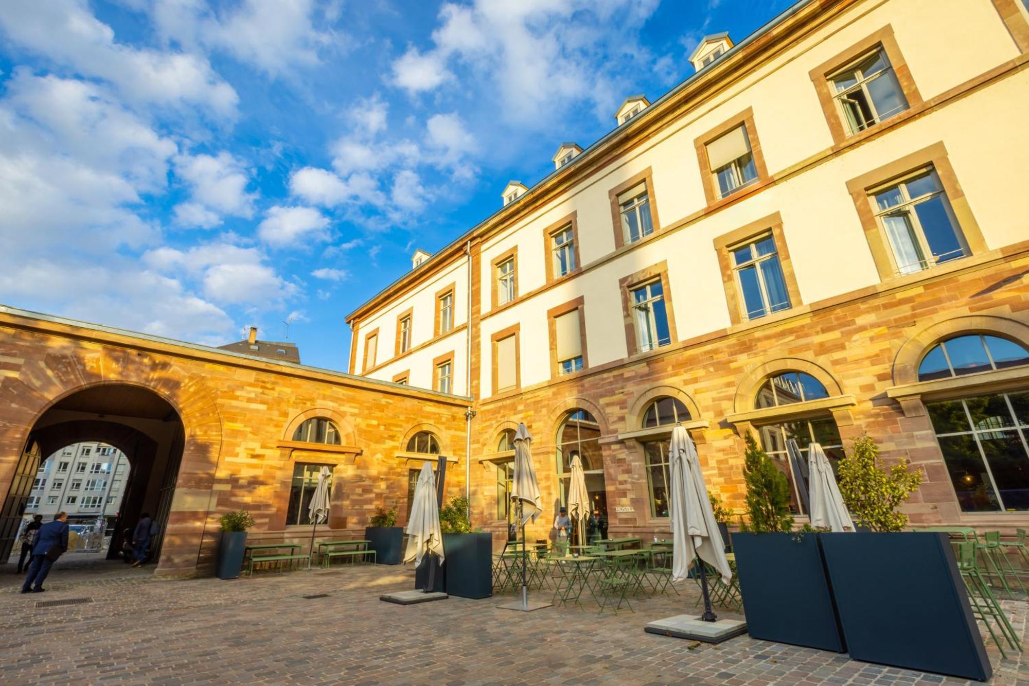 The People - Strasbourg Hostel Εξωτερικό φωτογραφία The photo shows a charming courtyard area with a historic building in the background. The architecture features warm-colored stone walls and large windows, suggesting a classic style. Sunlight illuminates the scene, highlighting the blue sky with sca