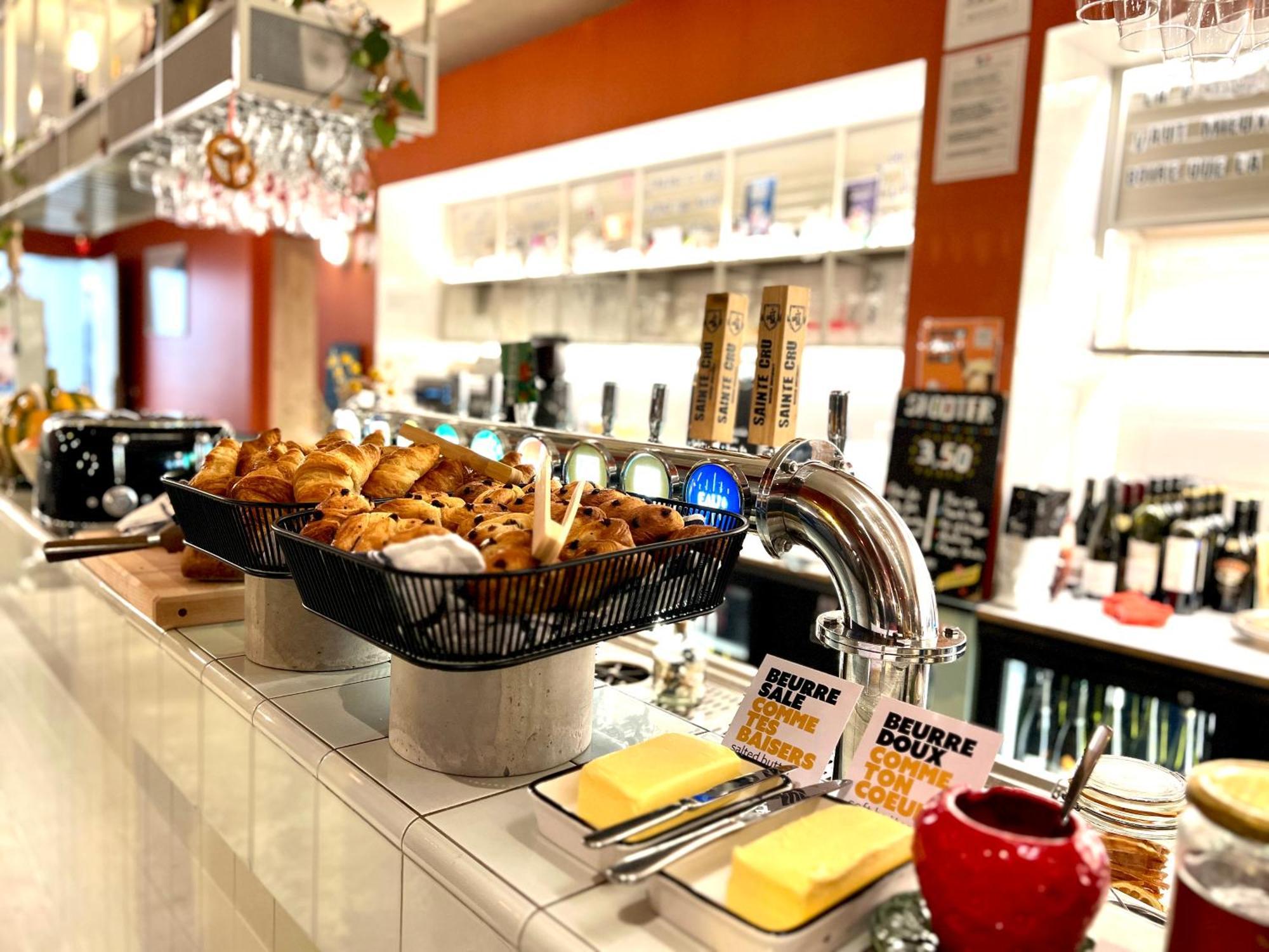 The People - Strasbourg Hostel Εξωτερικό φωτογραφία The photo shows a modern bar or café setup. In the foreground, there are baskets filled with various baked goods, likely pastries, suggesting a breakfast or snack option. To the right, there's a tap system, which might be for beverages like beer or s