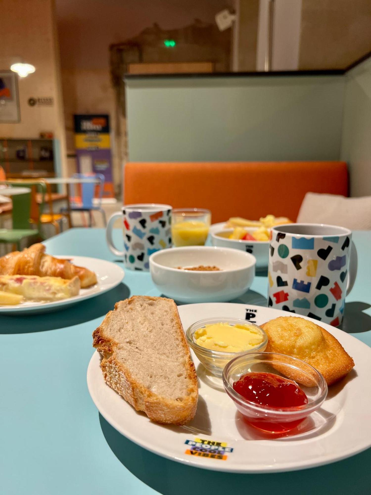 The People - Strasbourg Hostel Εξωτερικό φωτογραφία The photo shows a breakfast spread on a light blue table. It includes a plate with a slice of bread, a small pat of butter, a golden muffin, and containers of jam. There are also a few pastries, including croissants. In the background, there are two 